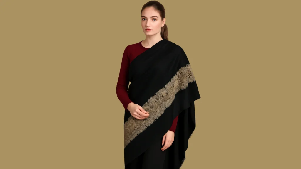 A woman wearing a traditional Kashmiri Pashmina shawl draped gracefully. The shawl is black with fine Sozni hand embroidery — intricate and delicate floral and paisley patterns stitched with thin silk threads in soft gold and ivory tones. The embroidery is flat and detailed, showing high craftsmanship.