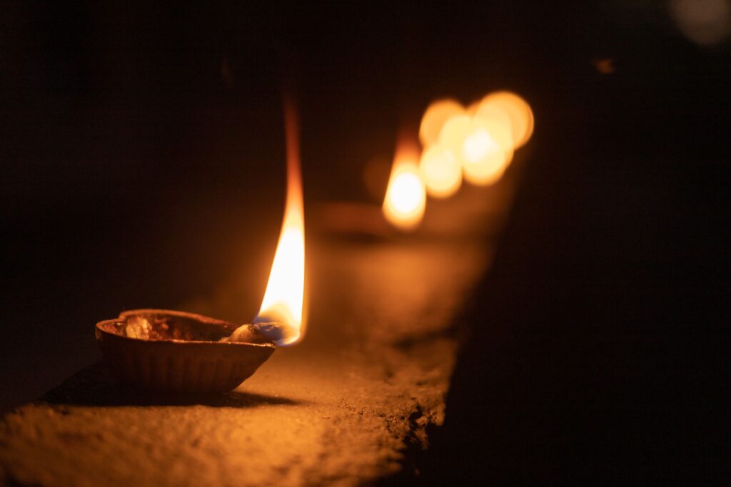 A clay diya oil lamp burning on a stone ledge, with a row of lamps out of focus behind it