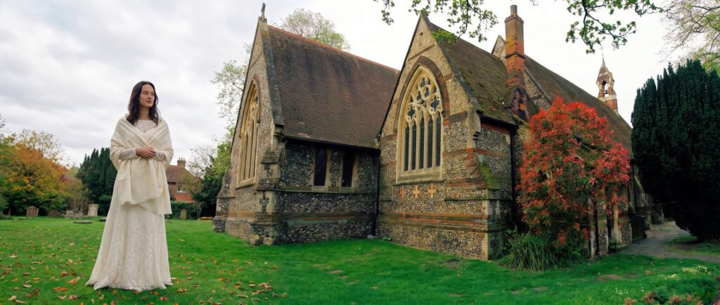 A bride in a lace gown and a cream pashmina on the lawn of a flint church with an autumn tree